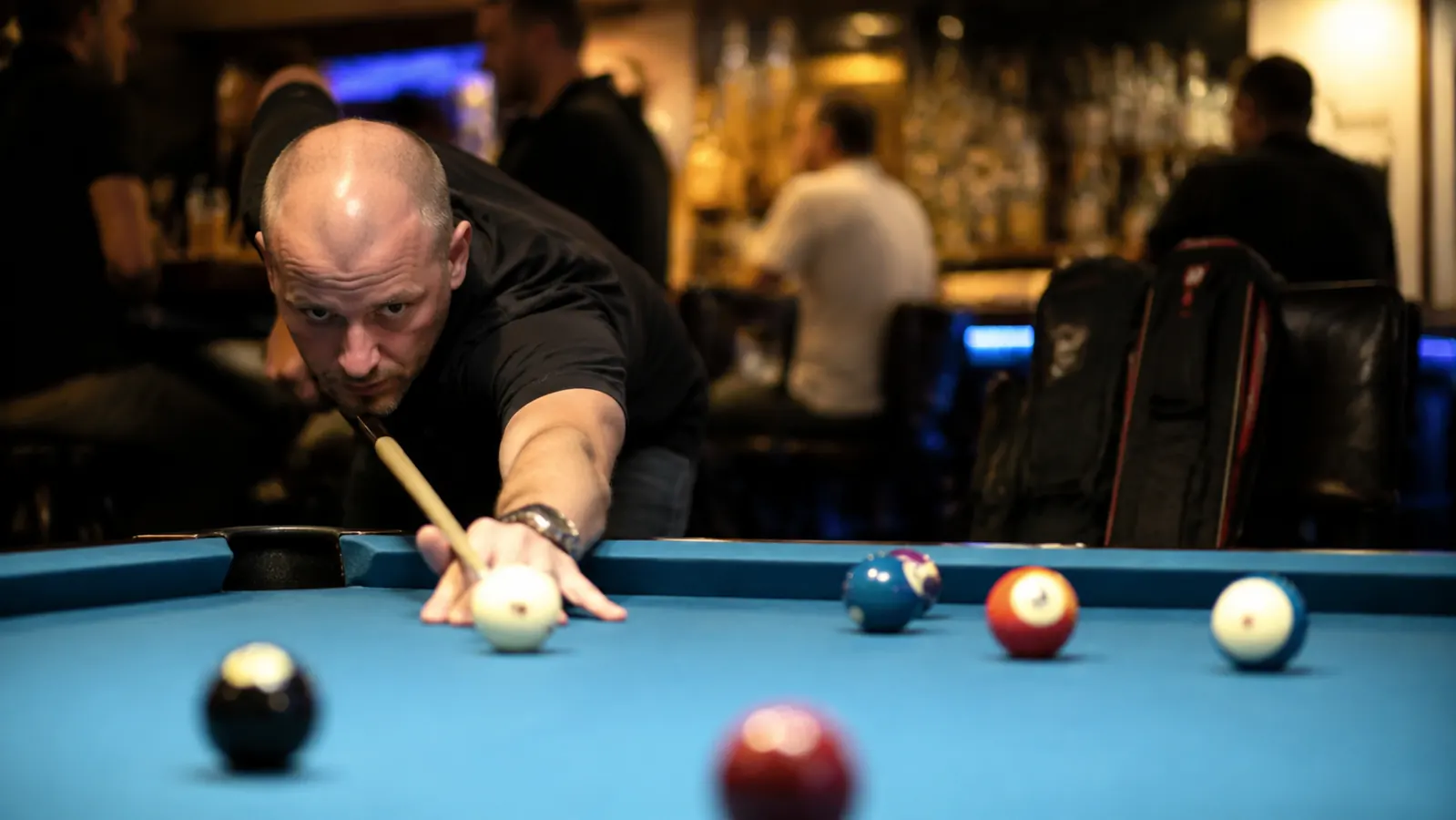 Pool player lining up a shot at a bar-style billiards table, personal cue case visible on a nearby chair