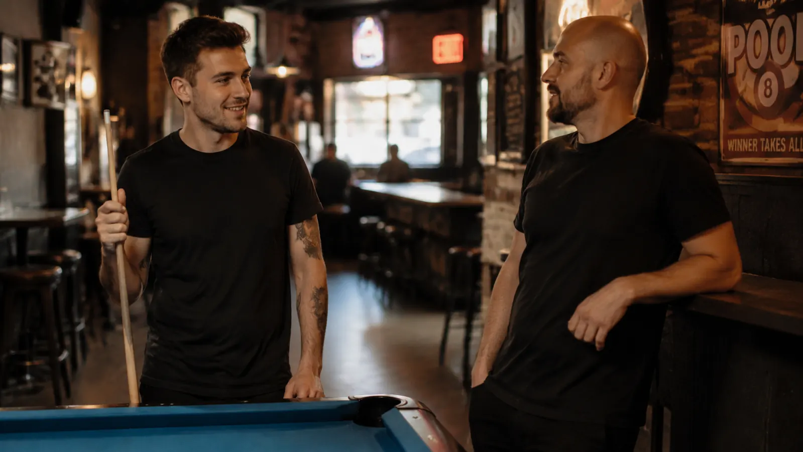 Pool player and teammate sharing a laugh at a league table, cue leaning against the rail