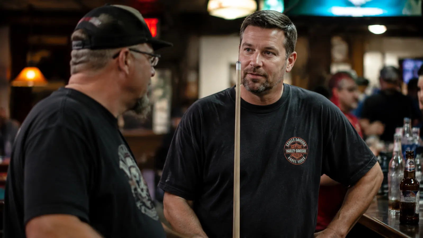 Pool player holding a break cue across his body in conversation at a bar, relaxed post-game pose